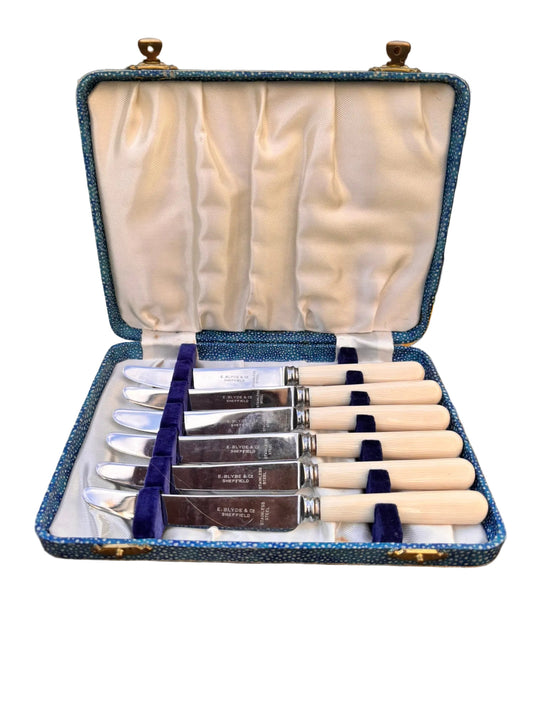 Set of butter knives in a decorative case on a floral-patterned surface with a blurred natural background.