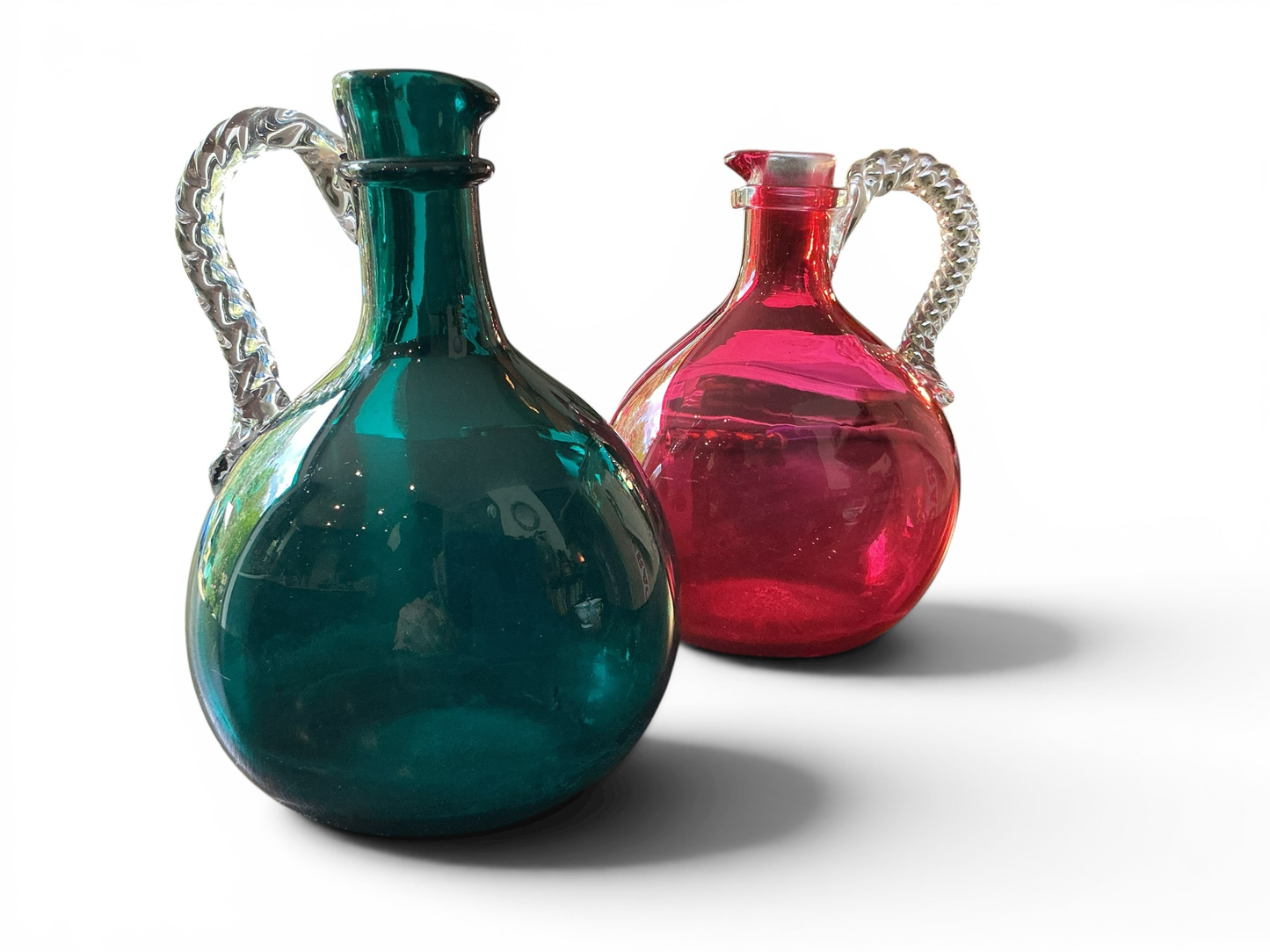 Otoch hand blown glass pitcher set in emerald green and ruby red with clear twisted handles and rounded silhouettes.