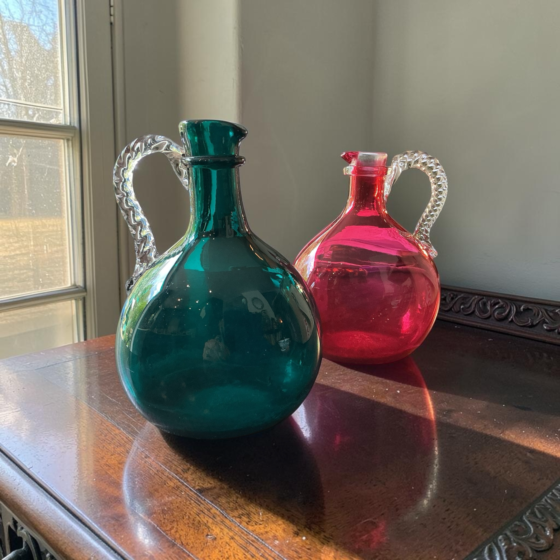 Otoch hand blown glass pitcher set in emerald green and ruby red with clear twisted handles on a wooden table.