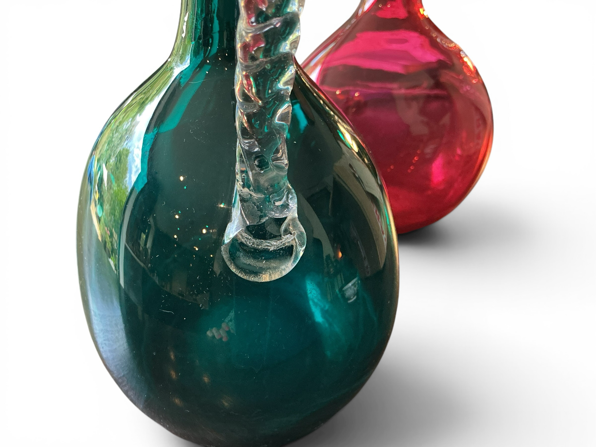 Otoch hand blown emerald green and ruby red glass pitchers with clear twisted handles on a white background