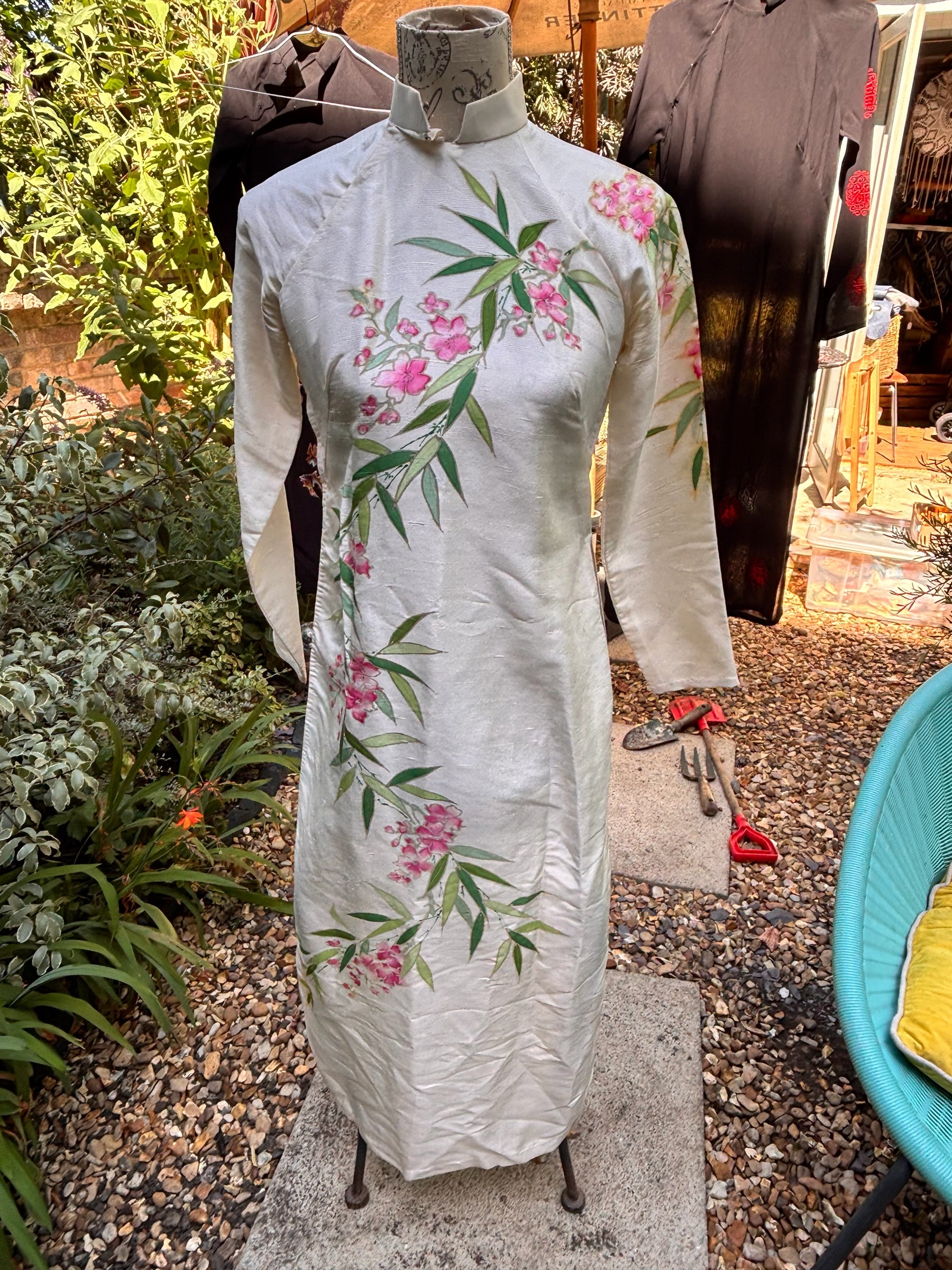Otoch off-white silk Ao Dai with hand-painted pink blossoms and green bamboo leaves on a mannequin in a garden.