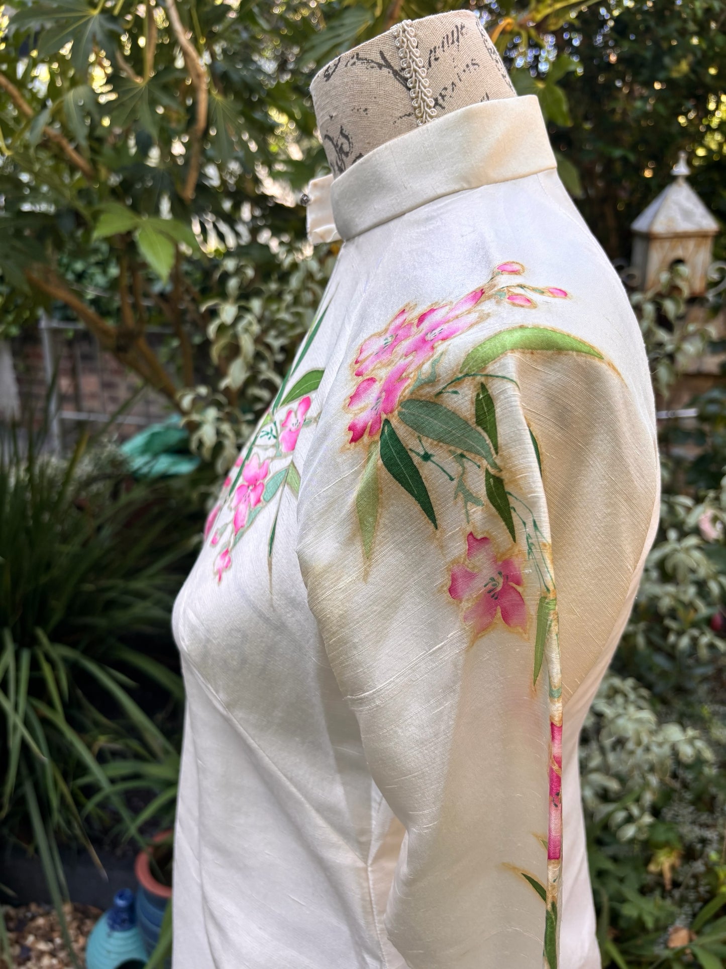 Otoch off-white silk Ao Dai featuring hand-painted pink floral blossoms and green bamboo leaves on a mandarin collar.