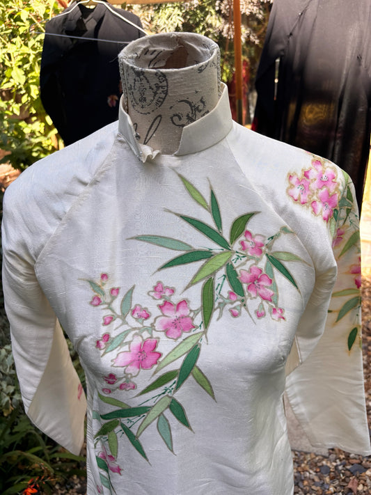Otoch off-white silk Ao Dai featuring hand-painted pink blossoms and green bamboo leaves on a mandarin collar tunic.